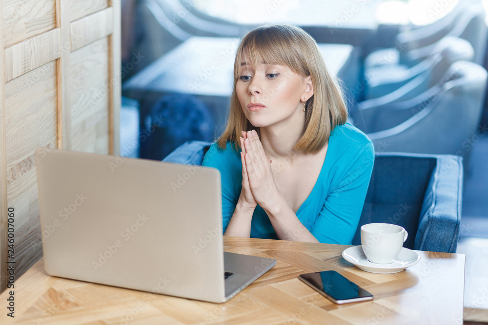 Fototapeta premium Please help me! Portrait of young beautiful student with blonde hair in blue t-shirt sitting in cafe and talking thought webcame with group mate begging for help with her homework