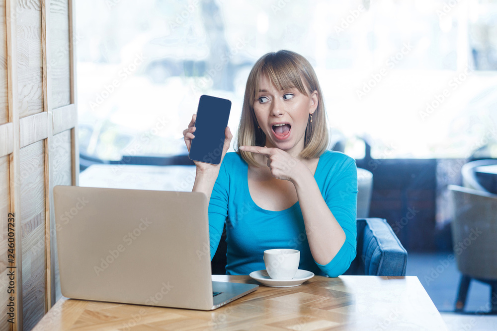Portrait of surprised beautiful young blonde woman in blue t-shirt, sitting with laptop, holding and pointing mobile display and looking with amazed. indoor, studio shot. Portrait of surprised beautiful young blonde woman in blue t-shirt, sitting with laptop, holding and pointing mobile display and looking with amazed. indoor, studio shot.