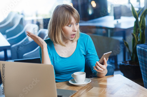 Top view portrait of young shocked blonde woman in blue t-shirt, sitting in cafe and holding her mobile smart phone and looking with amazed face and big eyes with raised hand. indoor studio shot. photo