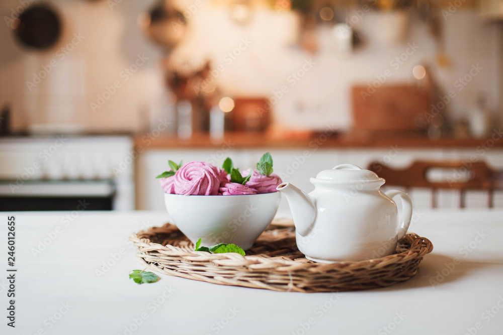 White teapot and pink dessert are serving on straw wicker tray on table. Tea time, cozy atmosphere hygge. Kitchen still life on background of warm light. Sweets and cake with herbal mint leaves.