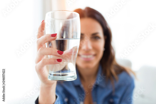 Pretty young woman smiling while looking at the camera through the glass of water at home.