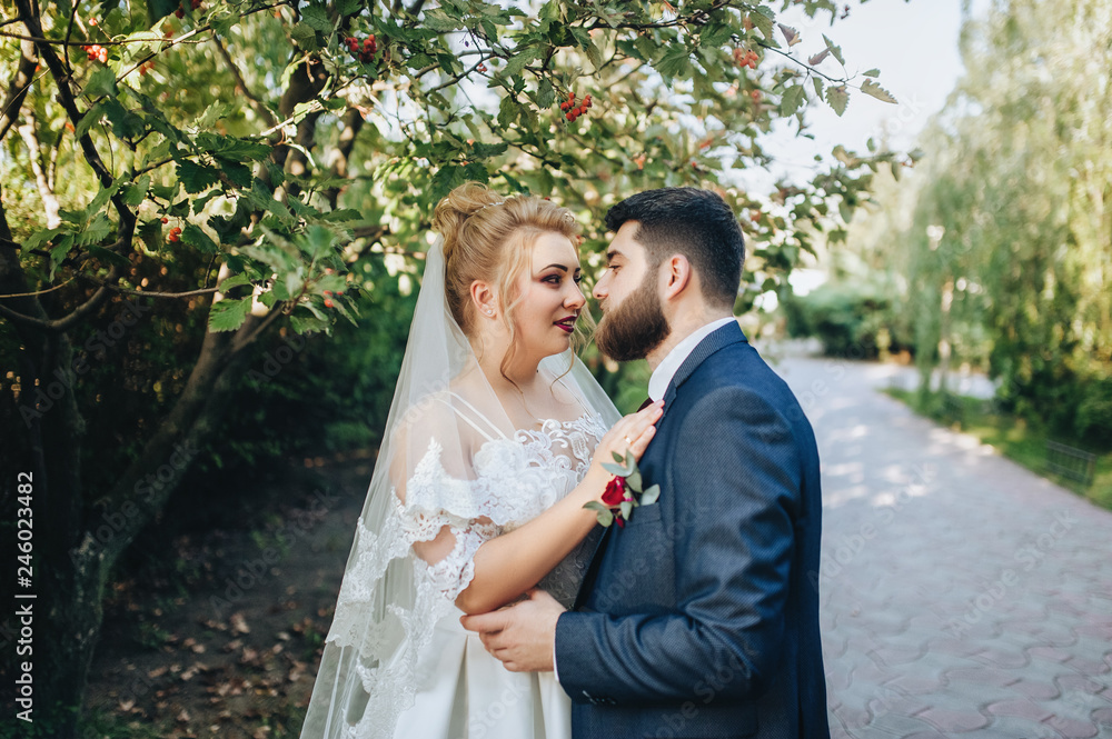 Bearded boyfriend in love and cute blonde bride hugging in a park on the nature. Portrait of lovers and smiling newlyweds in a green garden near the mountain ash. Wedding photography in foliage. Bearded boyfriend in love and cute blonde bride hugging in a park on the nature. Portrait of lovers and smiling newlyweds in a green garden near the mountain ash. Wedding photography in foliage.