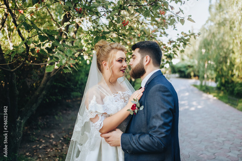 Bearded boyfriend in love and cute blonde bride hugging in a park on the nature. Portrait of lovers and smiling newlyweds in a green garden near the mountain ash. Wedding photography in foliage.