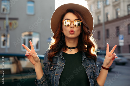 Fashion, beautiful brunette in brown hat, making duck face and showing peace by two hands. Attractive and stylish girl in sunglasses,choker, jeans jacket posing at street sending kiss at camera. photo