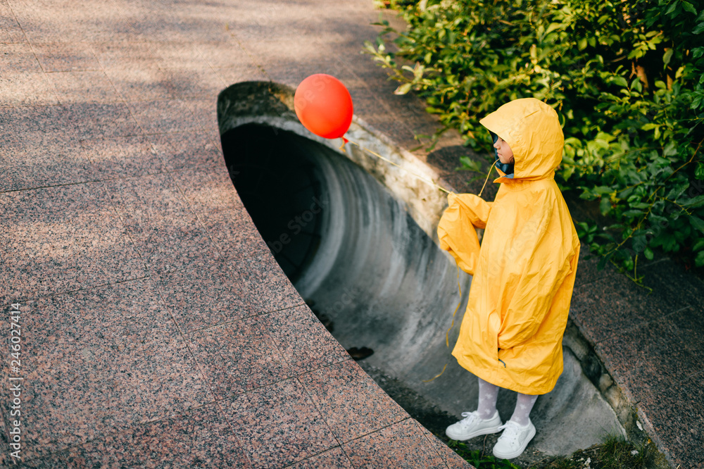 Portrait of pretty litle girl in big adult oversized yellow raincoat with red balloon in hand standing and sitting against sewage tunnel pipe. Scary movie concept. Funny child posing in summer day. - obrazy, fototapety, plakaty 