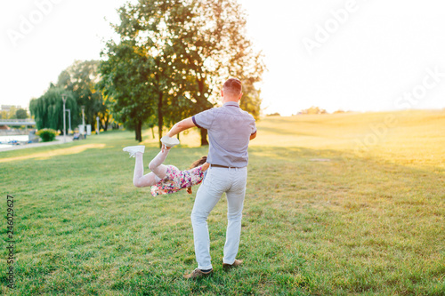 Handsome hipster father with mustache, hair style whirling his little daughter ootdoor on green field in park. Dad playing carousel with his cute child in multicolored dress. Parent with kid enjoying