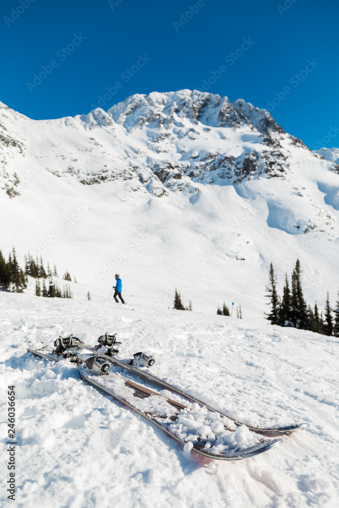 A pair of snow covered skis with the peak of Blackcomb and a skier in the background.