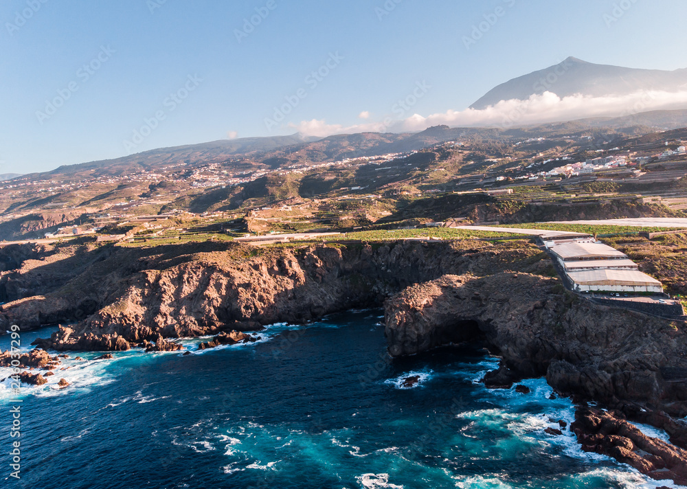 Cliff of volcano stones in the north of Tenerife with El Teide on the Top with clouds. Ocean and banana trees. Aerial picture with a drone 