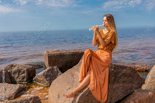 Young girl with long blond hair in long dress playing flute sitting on stone on sea shore