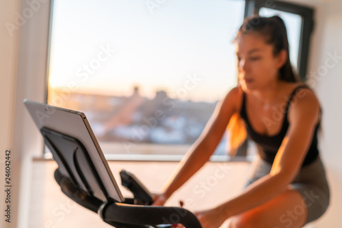 Home fitness fit woman exercising on smart stationary bike at home gym class watching screen online class biking exercise. Young girl training spinning the pedals pedaling. Focus on screen.