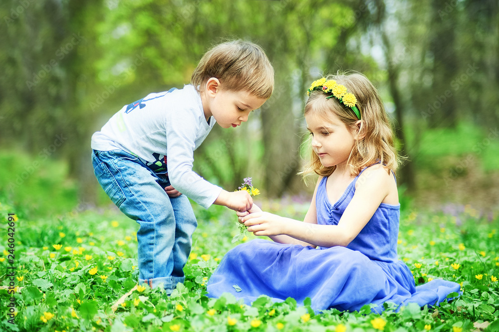 Fototapeta premium Brother and sister sit on the grass. Children's games, leisure. Two children are sitting on green meadow and smile. Boy and girl kiss on sunny day.