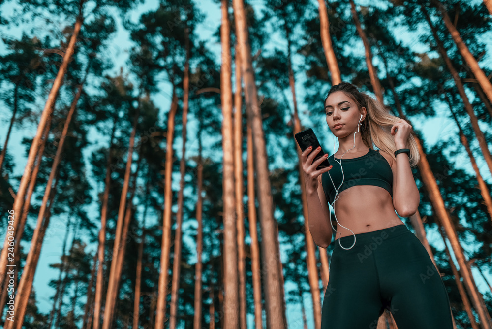 Beautiful girl athlete, stands in forest background of trees, in fresh air. Active lifestyle in park summer. Casual sportswear. In hands of phone and listens music on headphones. Free space for text.