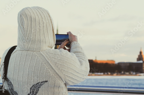 Man in white knitted jacket with hood taking photo of cityscape with his smartphone