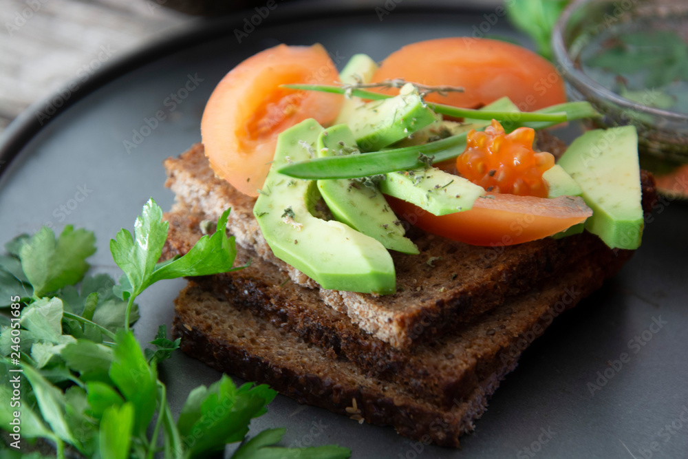 Healthy avocado sandwich on dark rye toast bread made with fresh sliced avocado, seeds and tomatoes on rustic wooden background.