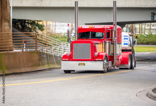 Big rig bright red classic American idol semi truck for long haul routs with high chrome exhaust pipes transporting semi trailer driving on the city street intersection turning under railroad bridge