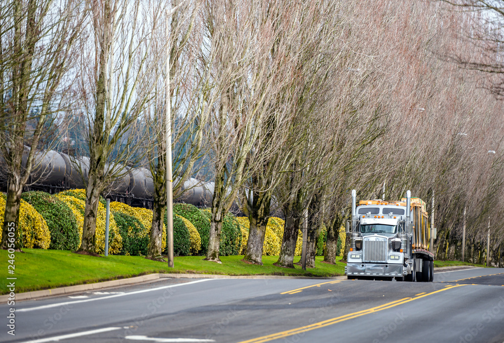 Classic big rig American semi truck with flat bed semi trailer transporting heavy equipment on the road with trees alley Classic big rig American semi truck with flat bed semi trailer transporting heavy equipment on the road with trees alley