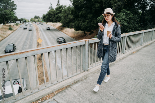 elegant office lady going to work in morning walking on pedestrian overpass outdoor using cellphone holding coffee. busy businesswoman crossing walkway bridge above the highway road car driving pass.