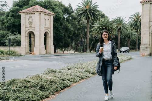 full length of young female exchange student going to take class walking in university. asian college girl holding coffee and carry bag in stanford. smart lady hard working at school going to course. photo