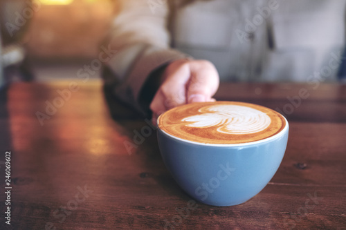 Closeup image of a hand holding a blue cup of hot latte coffee with latte art on wooden table in cafe