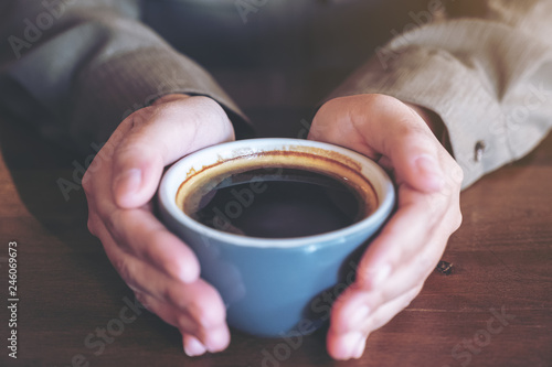 Closeup image of hands holding a blue cup of hot black coffee on wooden table in cafe