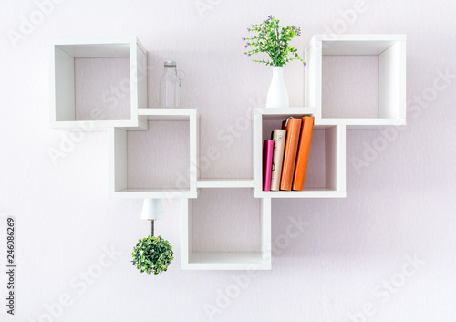 A modern white bookshelf on a white wall with a pair of books and a flowers. Minimalism style.