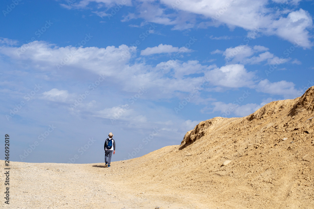 A boy hiker in a hat with a backpack is walking alone along a path in the Judean desert, mountain landscape against a blue sky