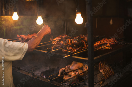 outdoor street chicken barbecue grill arm and hand brush oiling the grilling meat food broiler spit roasted over smoking living coals in a simple electric illumination, the Philippine lechon Manok