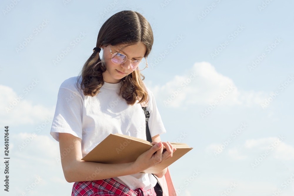 Outdoor portrait of teenager girl reading book. Girl student in glasses with backpack sky background with clouds Outdoor portrait of teenager girl reading book. Girl student in glasses with backpack sky background with clouds