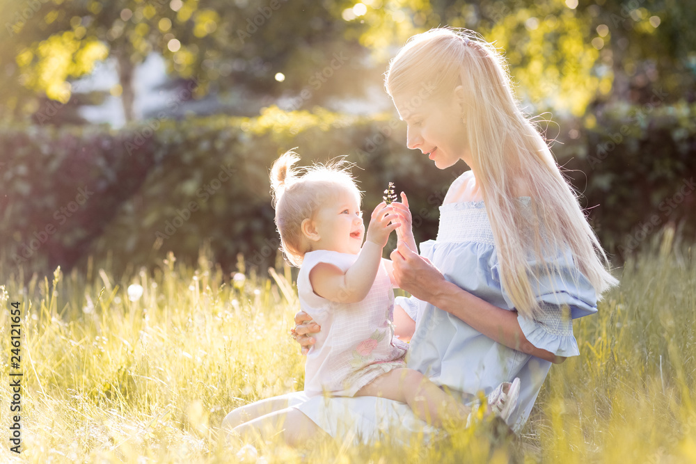 Young beautiful blonde mother with her baby girl laughing together and playing in green park outdoors at sunny day