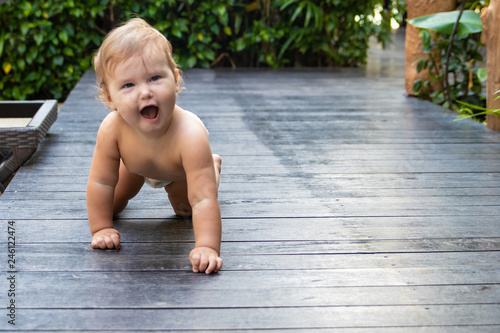 Pretty crawling baby girl smiling and crawling on the wooden brown walkway on the background of green bushes and trees. The child found on the floor and trying to eat. There is a place for text.
