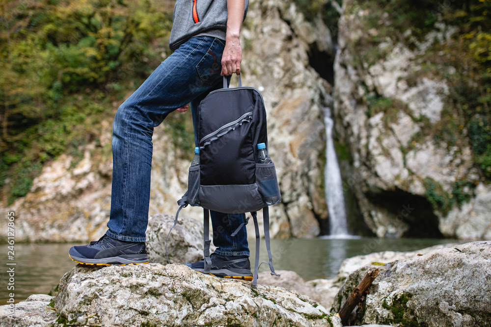 Unidentified man tourist in casual clothes is holding an equipped backpack for travel against the backdrop of a mountain river and stones. Concept of active recreation and comfortable clothing Unidentified man tourist in casual clothes is holding an equipped backpack for travel against the backdrop of a mountain river and stones. Concept of active recreation and comfortable clothing