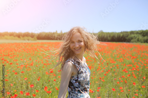 Beautiful girl is standing on summer field full of red poppy flowers in the grass. Sunny day with green lawn background. Happy woman in rustic dress is smiling. The wind in her hair.