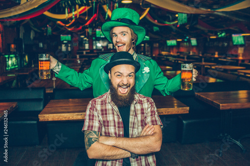 Happy and excited young men stand in pub and pose on camera. Guy in front keep hands crossed and smile. Young man behind wear green suit and has two mugs of beer.