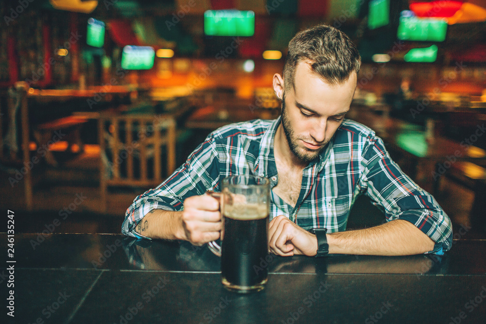 Young handsome man sitting alone at bar counter in pub. He hold mug of beer and look at watches.