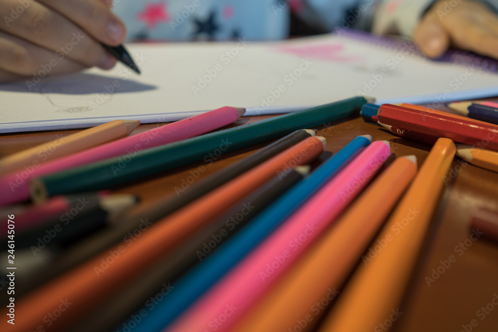 Colored pencils scattered on a wooden table. The child draws a picture in the album Colored pencils scattered on a wooden table. The child draws a picture in the album