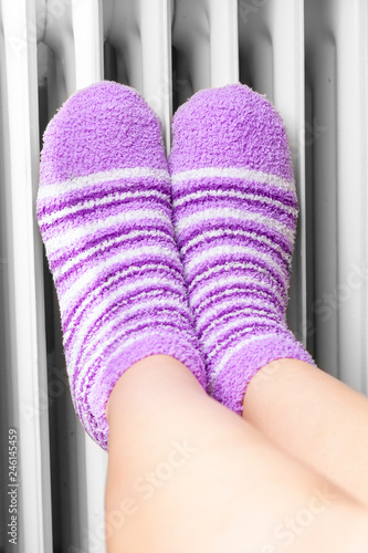  Woman in violet winter socks warm their feet near the heating radiator.