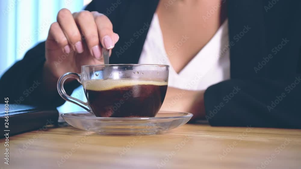 The girl is dressed in stylish business clothes sitting in the office and placing sugar in the coffee.