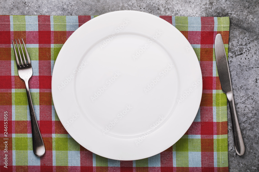 Empty ceramic plate of white color and cutlery on a checkered tablecloth, top view. Empty ceramic plate of white color and cutlery on a checkered tablecloth, top view.