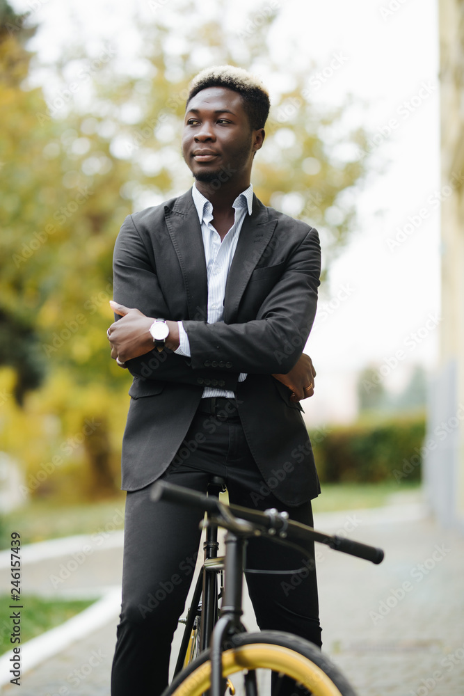 Handsome young Afro American man in suit leaning on his bike while standing on street outdoors Handsome young Afro American man in suit leaning on his bike while standing on street outdoors