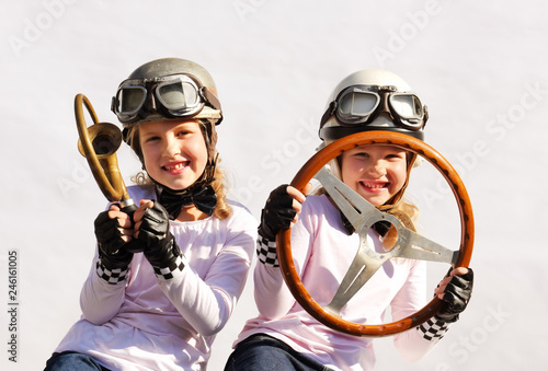 Twin sisters play race car driver sitting in their imaginative race car. They play happily wearing vintage helmets, goggles and use an old-timer steering wheel they found in the garage.