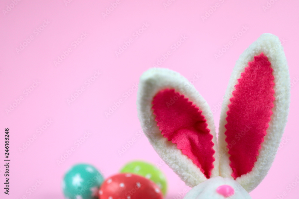 Fototapeta premium Easter background. White Easter bunny with red fur ears and colorful eggs on a pink background. Cropped shot, close-up, nobody, horizontal, blurred, free space. Easter concept.