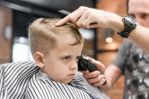 Hairdresser cuts hair little boy blonde with blue eyes making a fashionable hairstyle clipper close-up