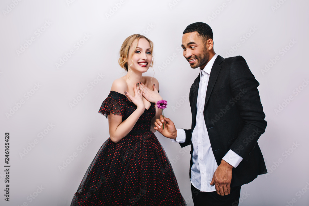 Happy cute couple in-love celebrating Valentine s day on white background. Attractive young blonde woman in luxury dress, handsome man in tuxedo, giving flower, smiling, positive emotions Happy cute couple in-love celebrating Valentine s day on white background. Attractive young blonde woman in luxury dress, handsome man in tuxedo, giving flower, smiling, positive emotions