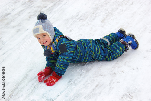 A four-year-old child, a boy, dressed in beautiful winter clothes, rides his stomach from a hill and laughs. look into the frame.