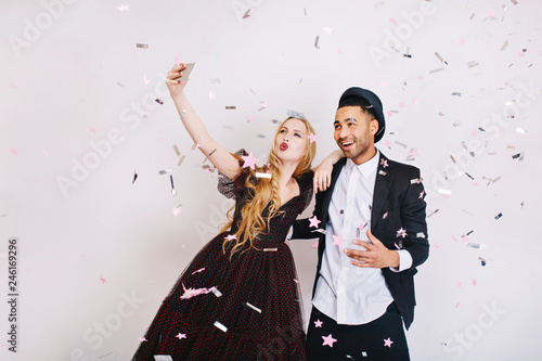 Crazy funny celebration of Valentine s day of couple in love in luxury evening clothes making selfie in tinsels on white background. Celebrating great party, smiling, expressing true positive emotions