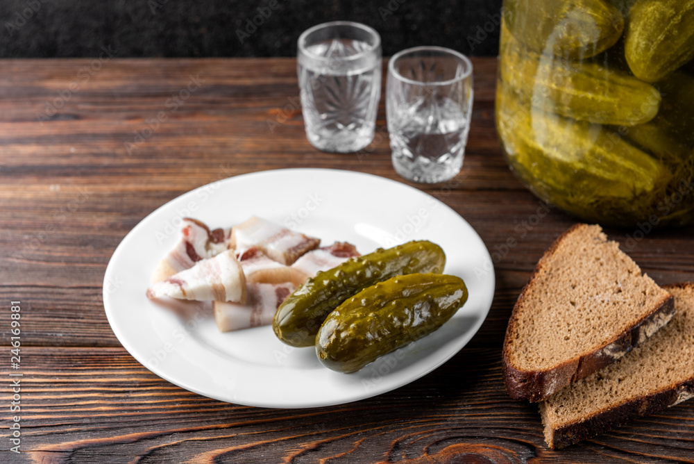 Vodka, salt pork fat, bread and pickled cucumbers on dark wooden background.