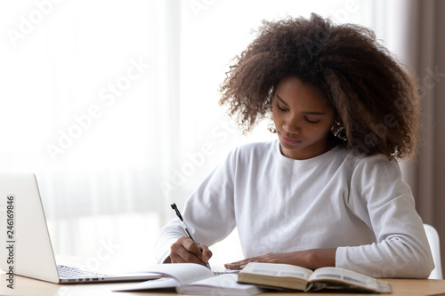 Focused African American teenage girl sit at table with laptop studying with handbooks, serious concentrated black teenager do homework at home, write down in notebook, using computer and textbook