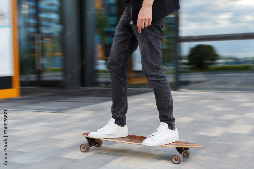 Shot of young man in black jeans and white sneakers riding a longboard in  urban area,