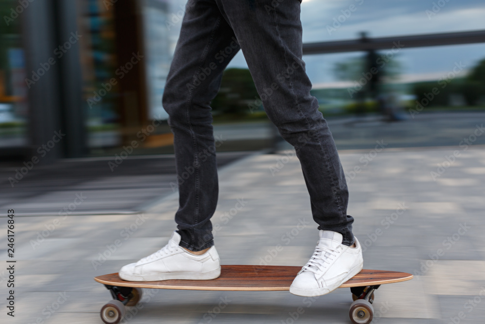Cropped shot of young man in black jeans and white sneakers riding on  longboard to the