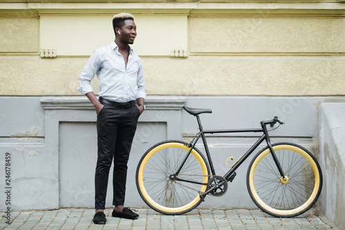 Handsome young Afro American man in casual clothes is using earphone, looking at camera and smiling while standing near his bike, standing outdoors